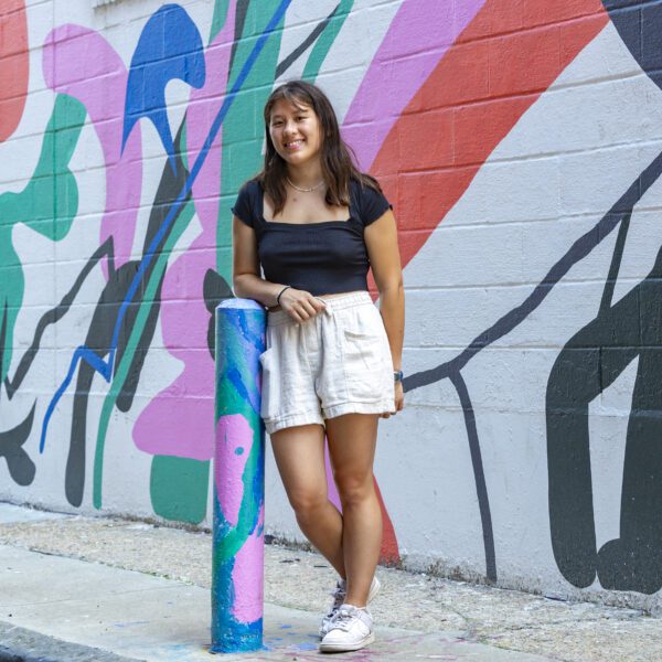 Kira Wu-Hacohen ’25 stands smiling in front of a colorful mural painted on a brick wall. She is wearing a black top, light shorts, and white sneakers, and is leaning casually against a painted post on a city sidewalk.