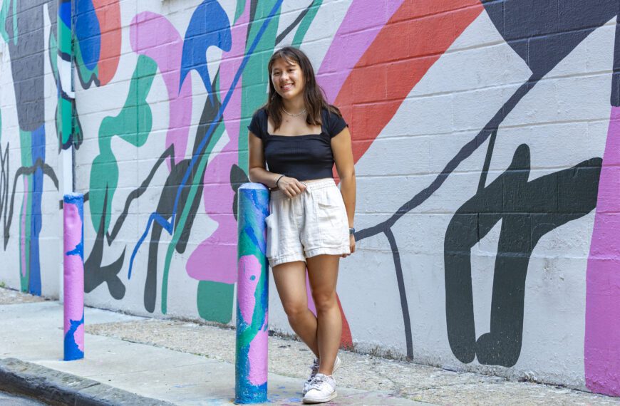 Kira Wu-Hacohen ’25 stands smiling in front of a colorful mural painted on a brick wall. She is wearing a black top, light shorts, and white sneakers, and is leaning casually against a painted post on a city sidewalk.