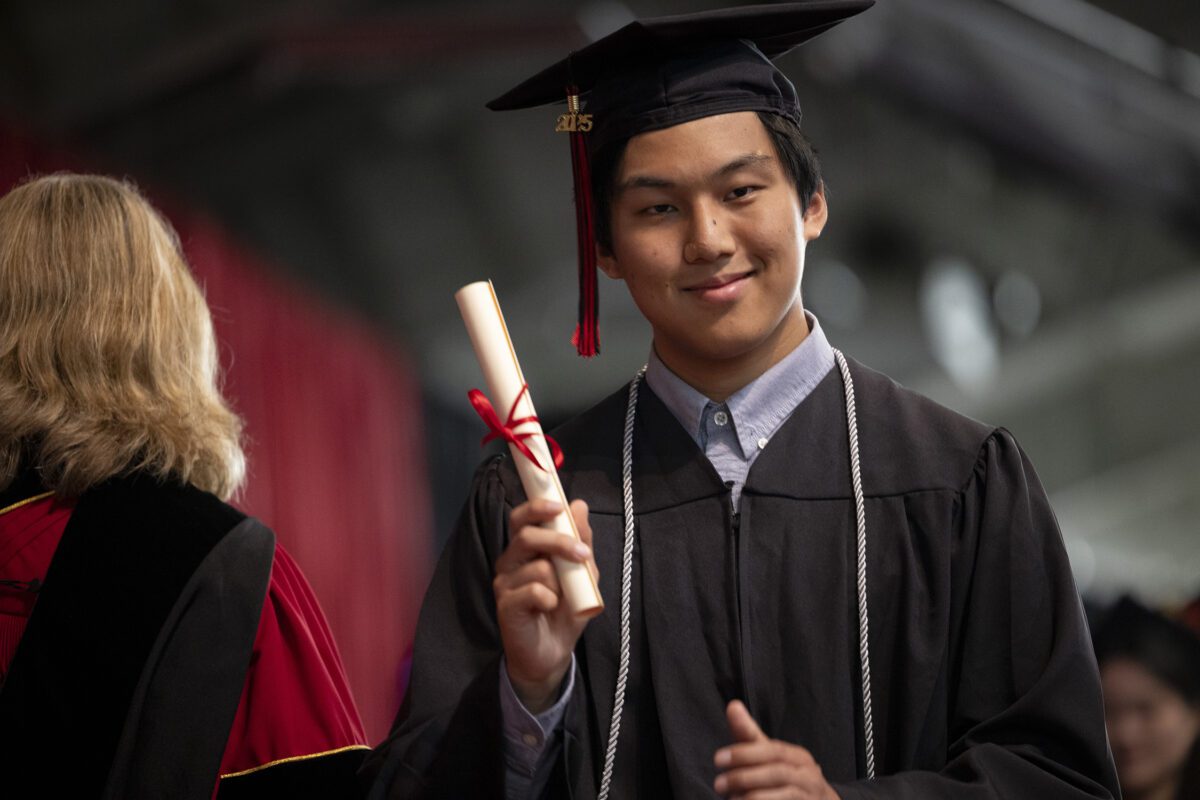 Kenneth Hsu ’25 smiles while holding his diploma during Haverford College’s Commencement ceremony. He is wearing a graduation cap with a red and black tassel, a black gown, and an honor cord.