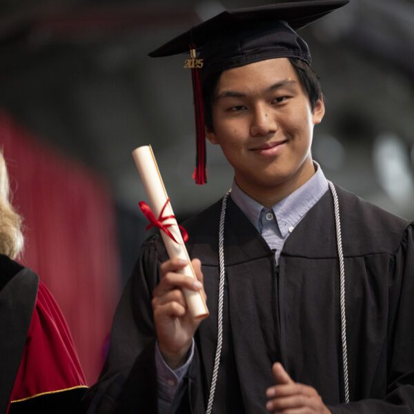 Kenneth Hsu ’25 smiles while holding his diploma during Haverford College’s Commencement ceremony. He is wearing a graduation cap with a red and black tassel, a black gown, and an honor cord.