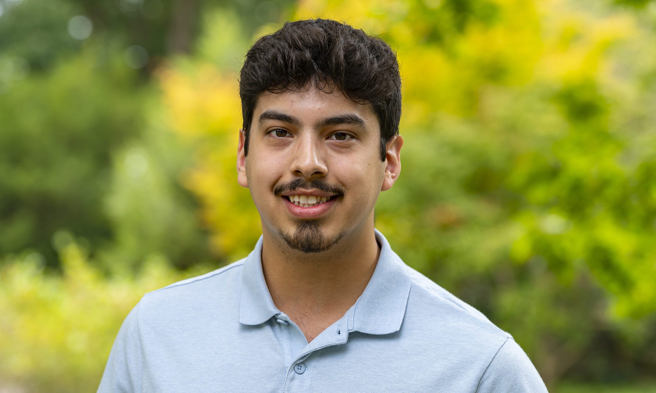 A young man wearing a light blue polo shirt smiles at the camera while standing outdoors. The background is filled with vibrant green and yellow foliage, softly blurred to highlight the subject.