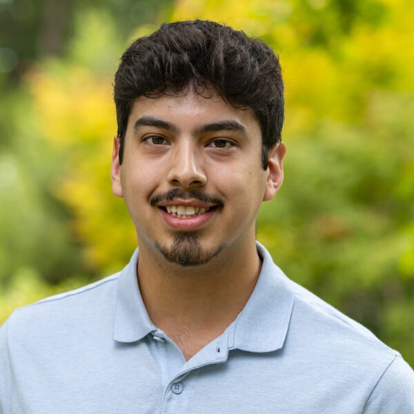 A young man wearing a light blue polo shirt smiles at the camera while standing outdoors. The background is filled with vibrant green and yellow foliage, softly blurred to highlight the subject.