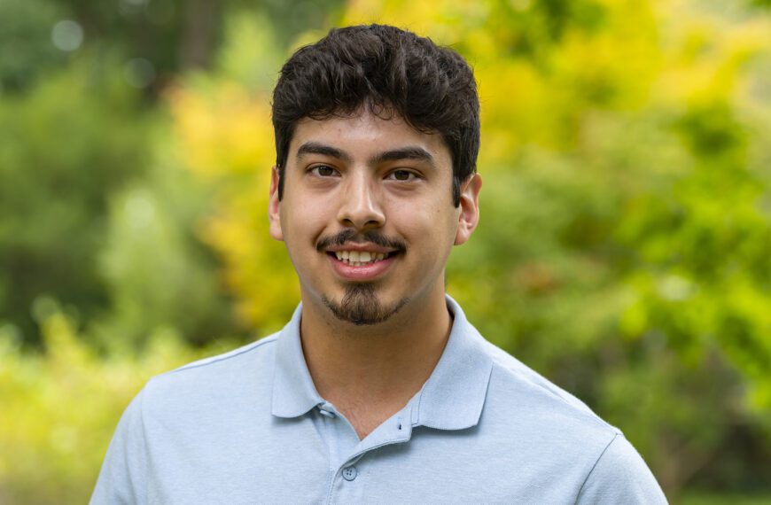 A young man wearing a light blue polo shirt smiles at the camera while standing outdoors. The background is filled with vibrant green and yellow foliage, softly blurred to highlight the subject.