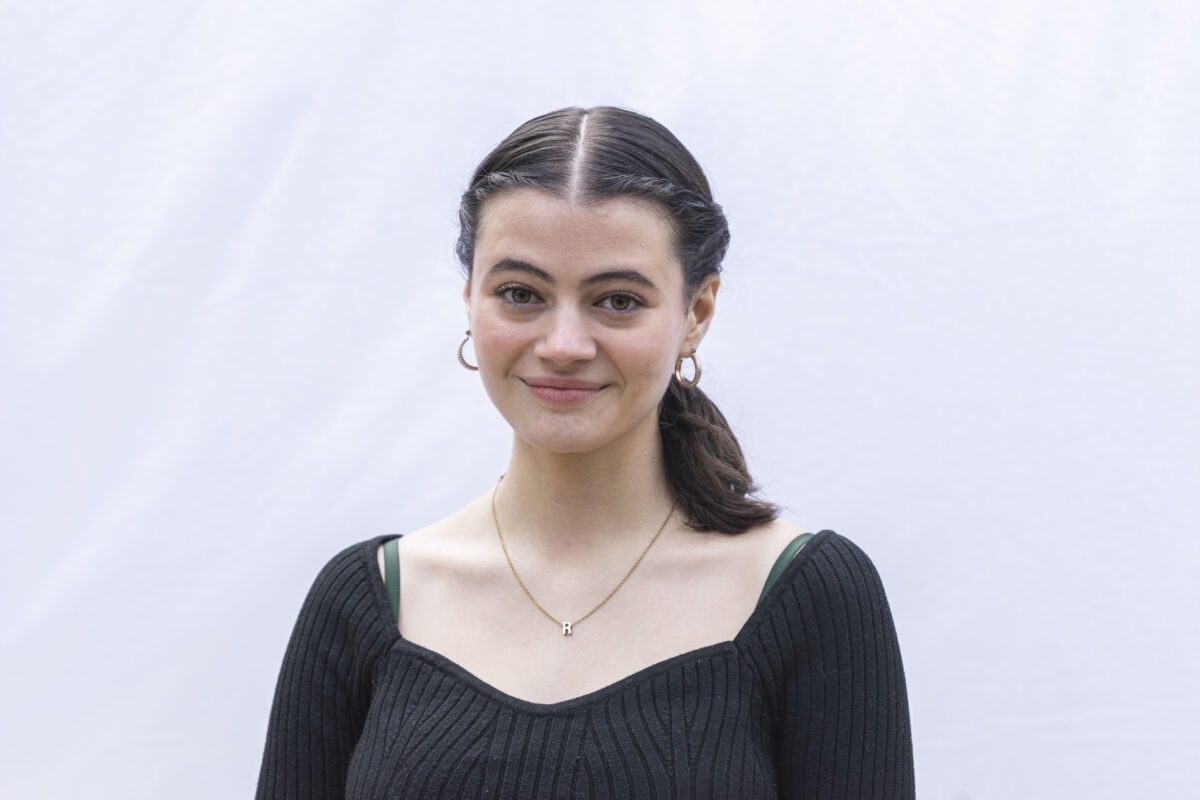 A young woman with dark hair pulled back in a ponytail smiles slightly while standing in front of a light-colored backdrop. She is wearing a black ribbed top, gold hoop earrings, and a necklace with a small letter "R" pendant.