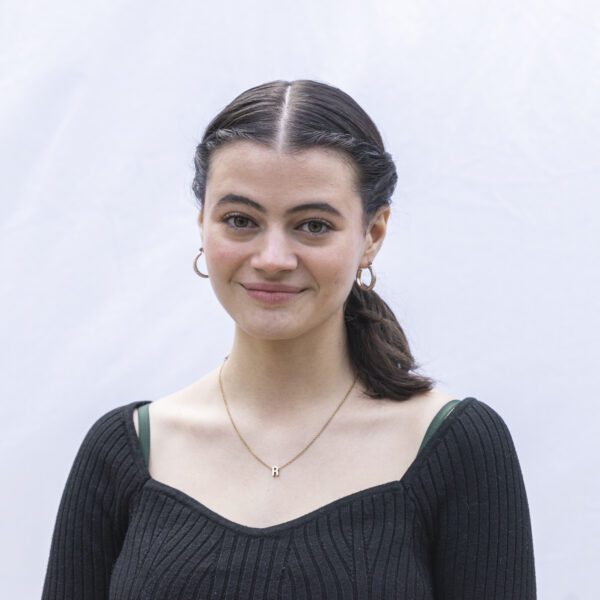 A young woman with dark hair pulled back in a ponytail smiles slightly while standing in front of a light-colored backdrop. She is wearing a black ribbed top, gold hoop earrings, and a necklace with a small letter "R" pendant.