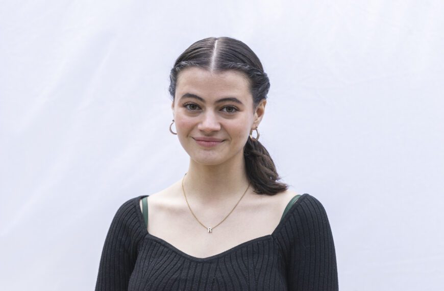 A young woman with dark hair pulled back in a ponytail smiles slightly while standing in front of a light-colored backdrop. She is wearing a black ribbed top, gold hoop earrings, and a necklace with a small letter "R" pendant.