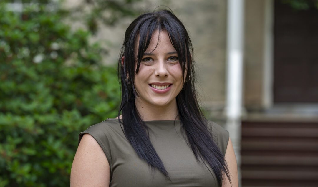 A young woman with long dark hair and bangs smiles at the camera while standing outdoors. She is wearing a sleeveless olive green top, and the background features greenery and the steps of a building.