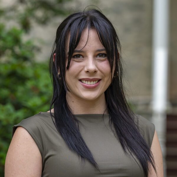 A young woman with long dark hair and bangs smiles at the camera while standing outdoors. She is wearing a sleeveless olive green top, and the background features greenery and the steps of a building.