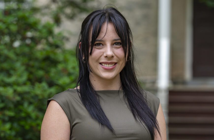 A young woman with long dark hair and bangs smiles at the camera while standing outdoors. She is wearing a sleeveless olive green top, and the background features greenery and the steps of a building.