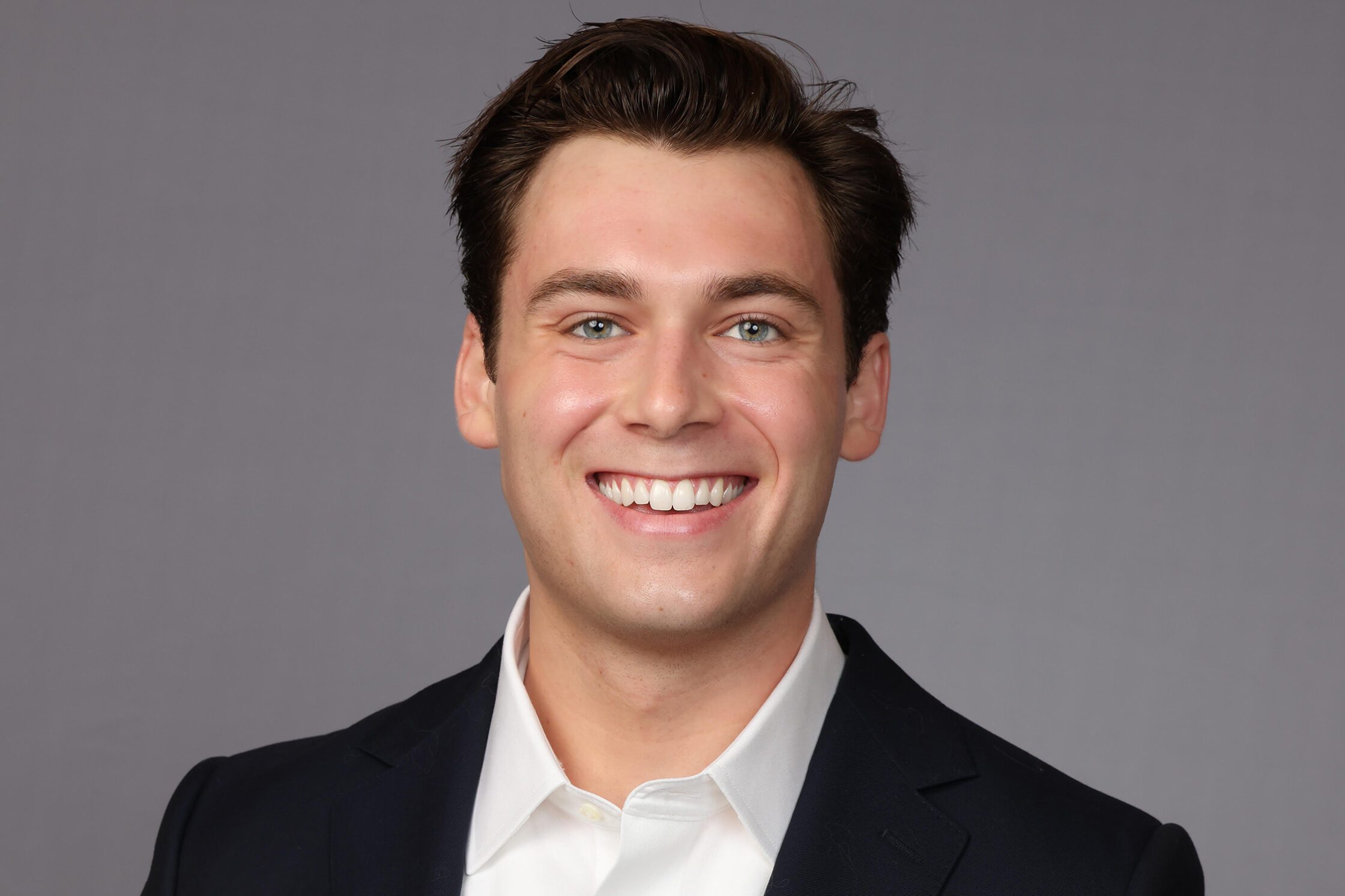 A young man with short dark hair smiles at the camera while wearing a dark suit jacket and a white collared shirt. The background is a plain gray color.