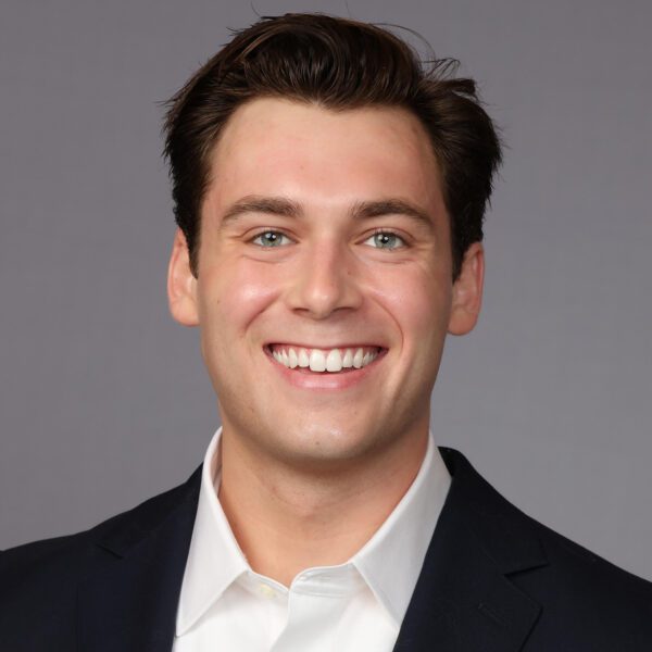 A young man with short dark hair smiles at the camera while wearing a dark suit jacket and a white collared shirt. The background is a plain gray color.