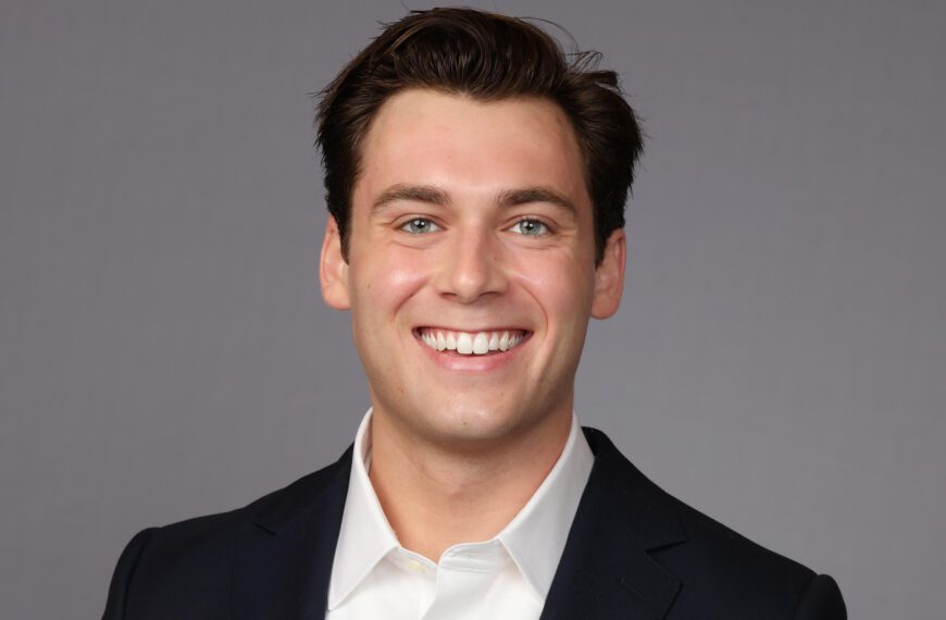 A young man with short dark hair smiles at the camera while wearing a dark suit jacket and a white collared shirt. The background is a plain gray color.