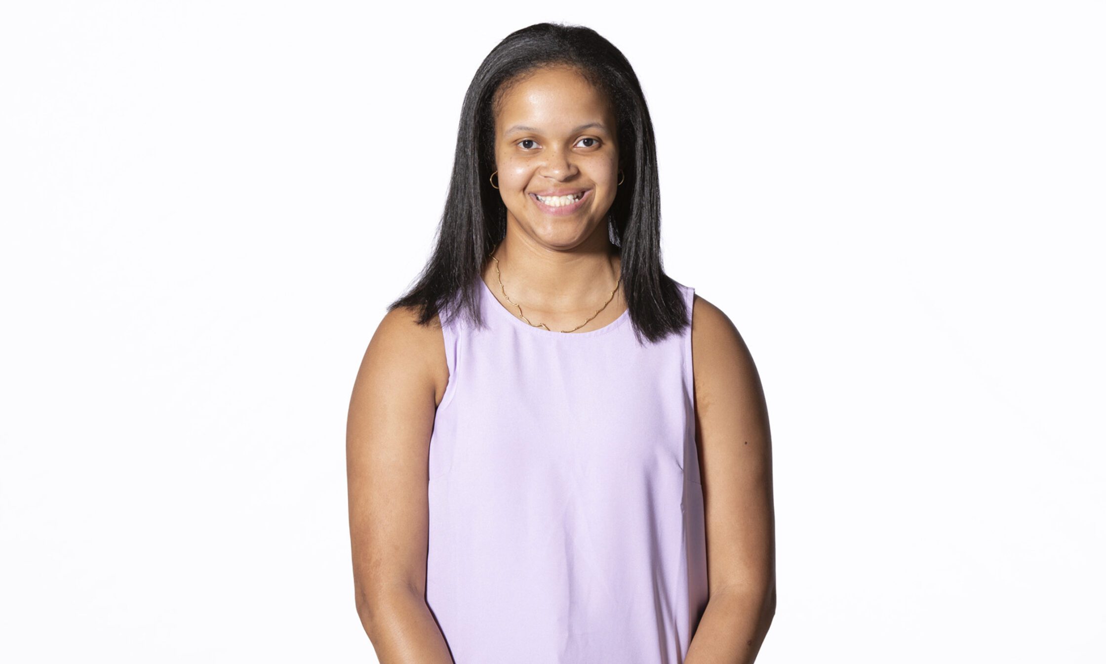 A young woman with straight dark hair smiles at the camera while wearing a sleeveless light purple top, gold hoop earrings, and a delicate gold necklace. The background is plain white.