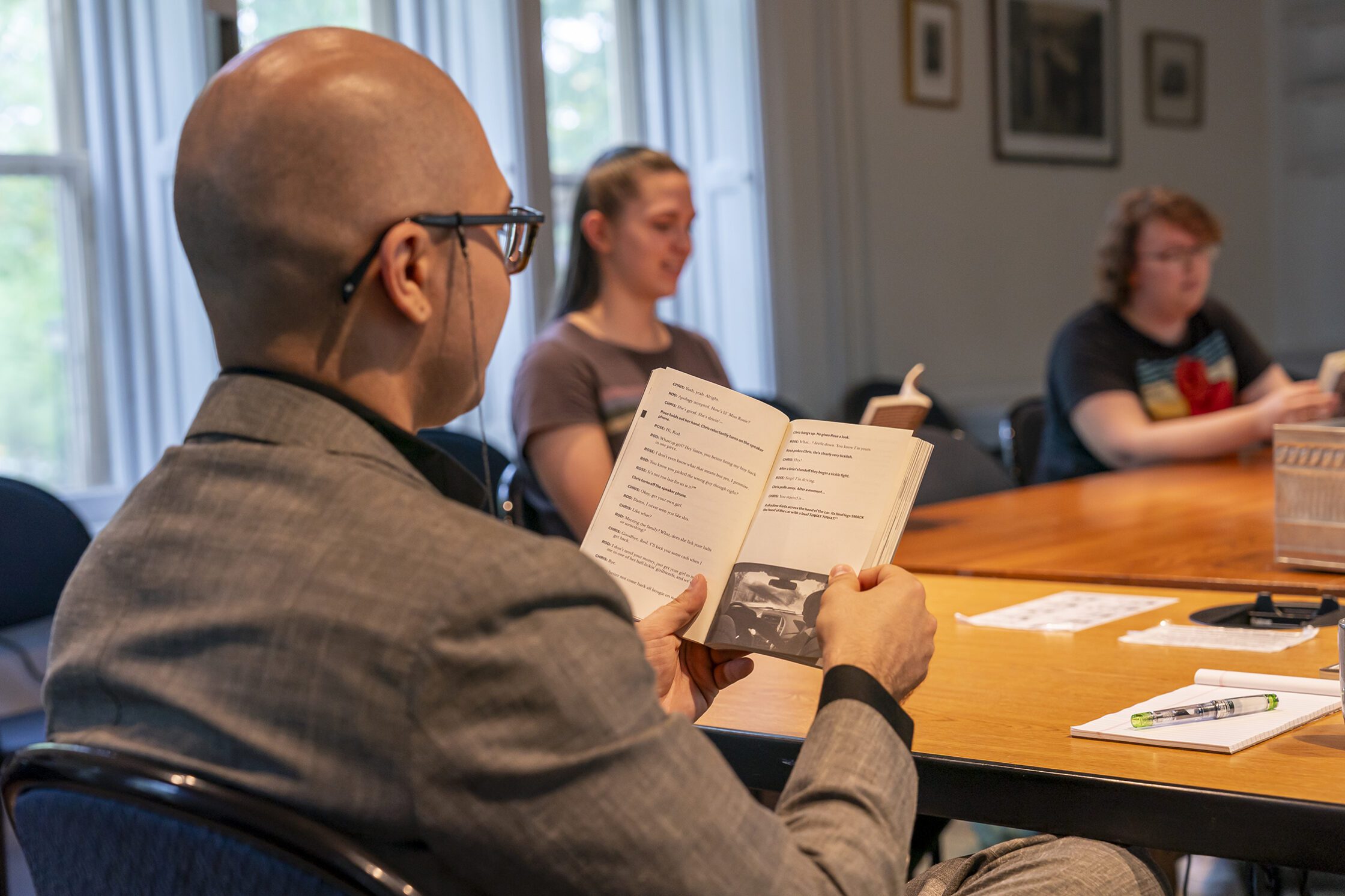 A person in a gray suit reads from an open book during a classroom discussion, seated at a wooden table with a pen and notebook nearby. Two other participants sit farther down the table, also reading, in a room lit by large windows.