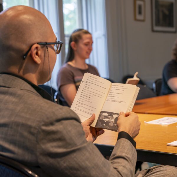 A person in a gray suit reads from an open book during a classroom discussion, seated at a wooden table with a pen and notebook nearby. Two other participants sit farther down the table, also reading, in a room lit by large windows.