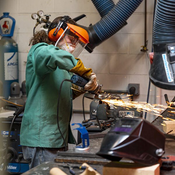 A student wearing a green shop coat, gloves, and a face shield uses a handheld grinder on a metal piece secured in a vise, with bright sparks flying in a workshop setting.