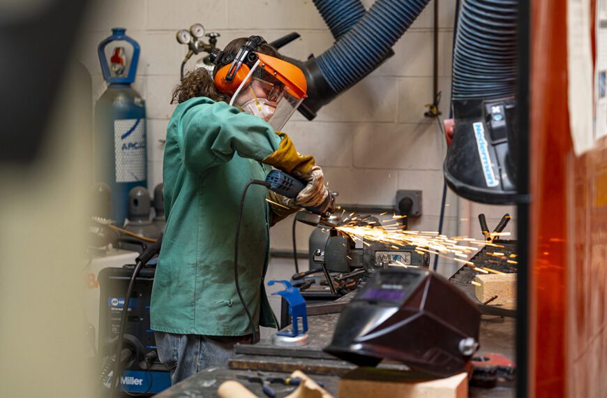 A student wearing a green shop coat, gloves, and a face shield uses a handheld grinder on a metal piece secured in a vise, with bright sparks flying in a workshop setting.