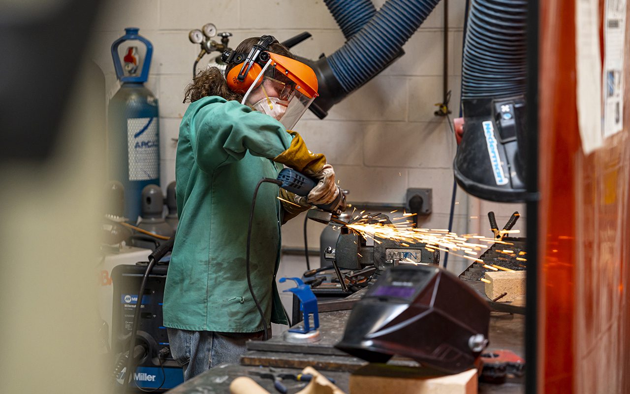 A student wearing a green shop coat, gloves, and a face shield uses a handheld grinder on a metal piece secured in a vise, with bright sparks flying in a workshop setting.