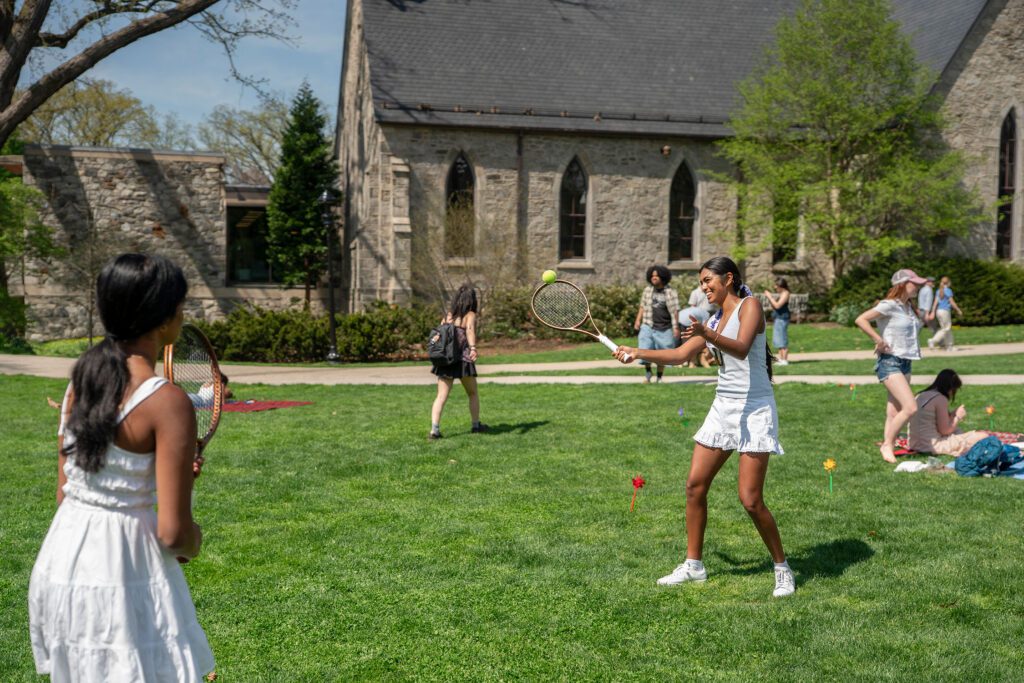 Two students in white outfits play tennis on the lawn during Pinwheel Day, with Founders Hall and other students relaxing and walking in the background.
