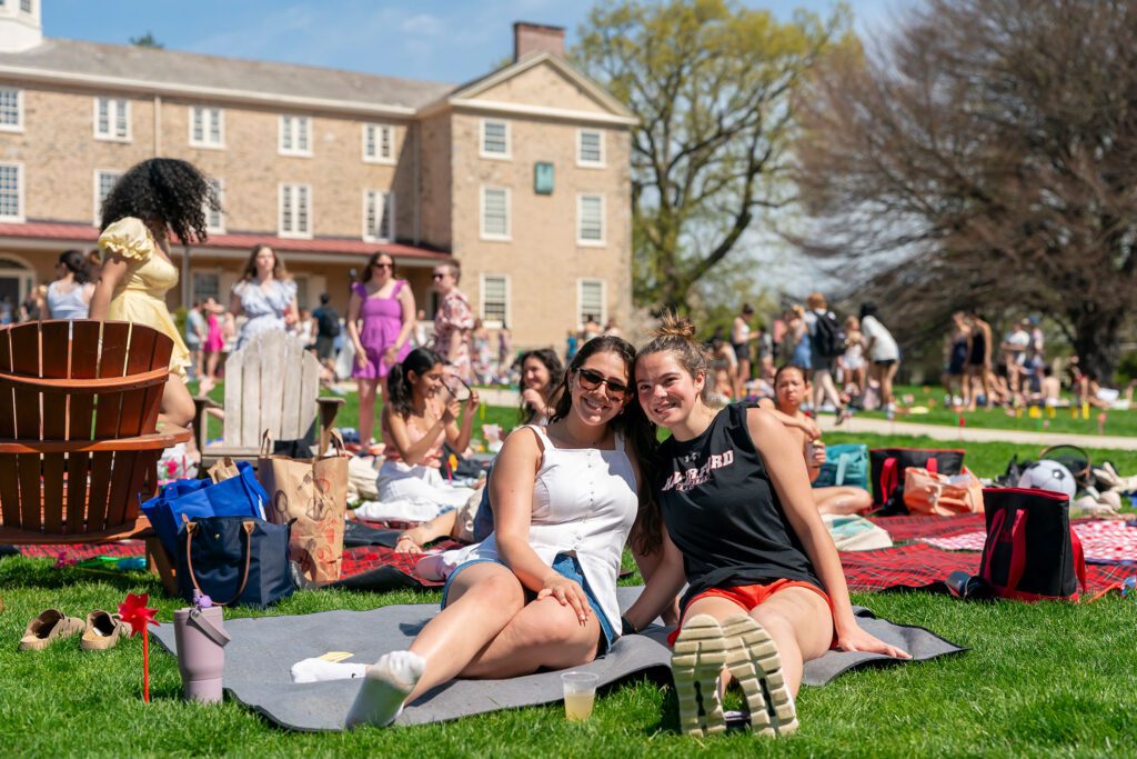 Two students sit close together on a blanket and smile for the camera as groups of classmates gather across the sunny lawn behind them.