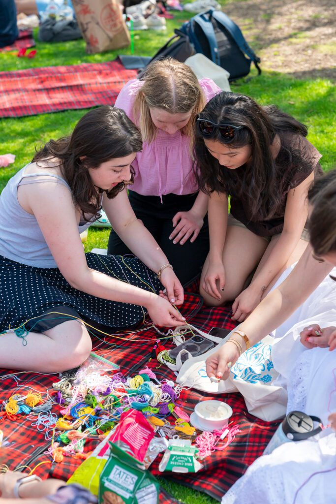 Three students sit on a plaid blanket working on a craft project, surrounded by colorful embroidery floss, snacks, and bags on the grass.