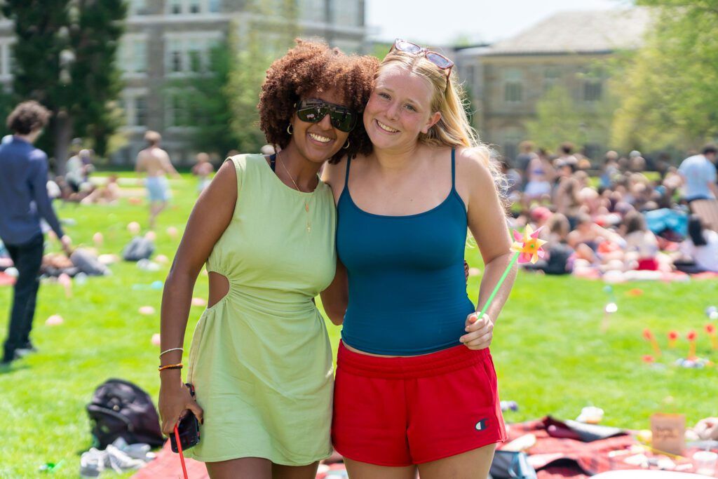 Two students stand arm in arm on the lawn, smiling for a photo while one holds a colorful pinwheel and other Pinwheel Day festivities continue behind them.