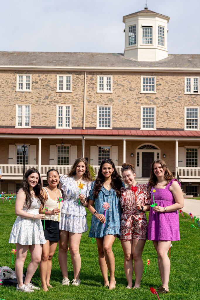 Five students stand in front of Founders Hall holding colorful pinwheels and smiling for a group photo on the lawn.