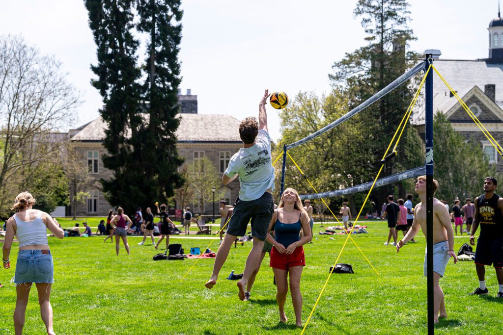 A student jumps to hit a volleyball over the net while classmates watch and play on the lawn in front of campus buildings.