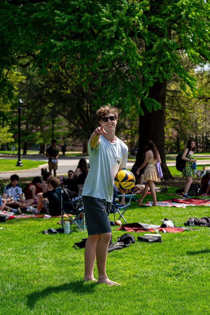 A barefoot student wearing sunglasses stands on the grass holding a volleyball and pointing toward the camera, with other students gathered behind him.