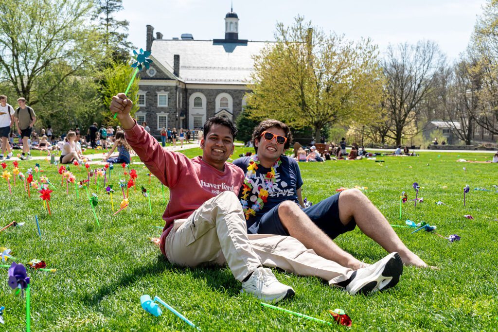 Two students sit on the grass among colorful pinwheels, smiling at the camera as one raises a blue pinwheel.