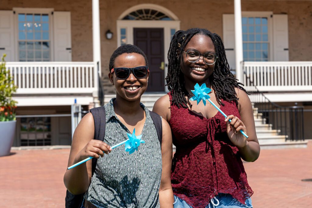 Two students stand smiling in front of Founders Hall, each holding a blue pinwheel.