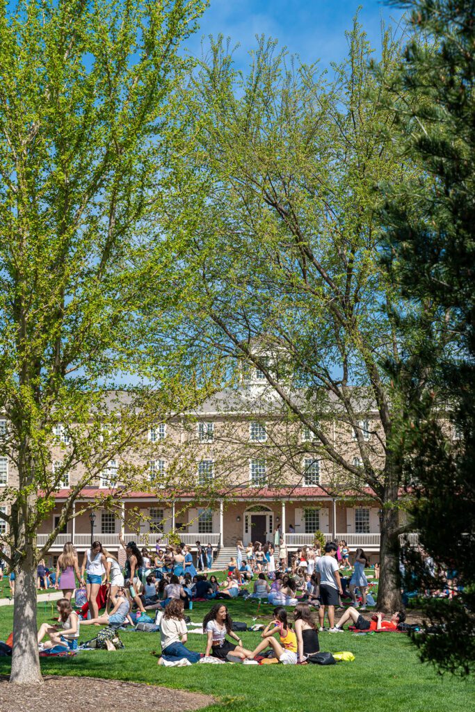 Students lounge and talk on the lawn in front of Founders Hall, framed by leafy spring trees and scattered pinwheels.