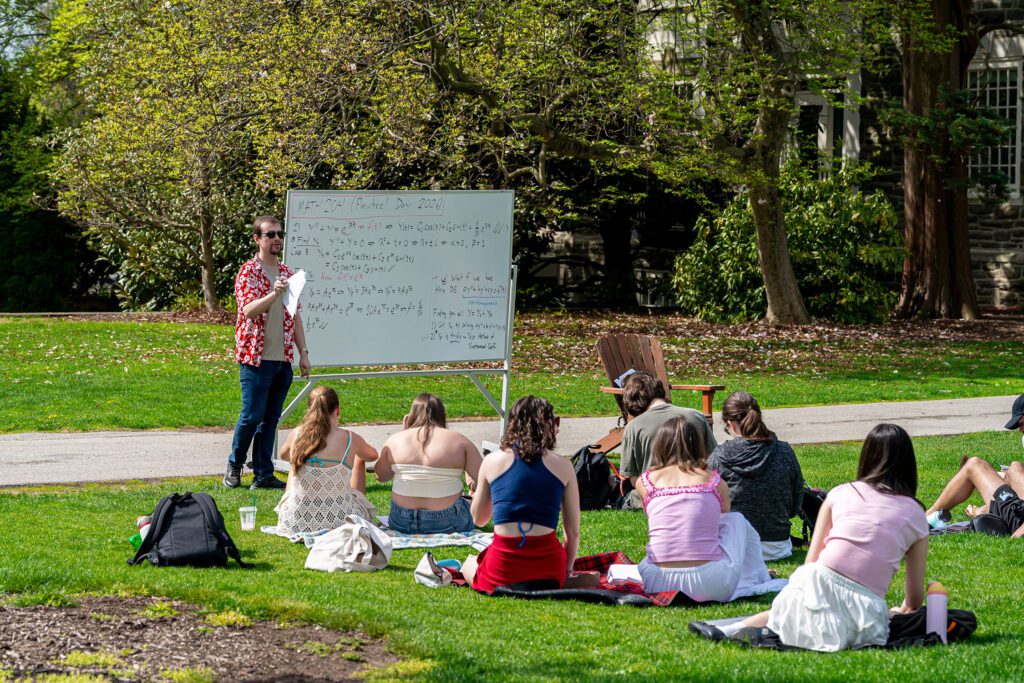 A professor stands beside a whiteboard filled with math equations, teaching an outdoor class to students seated on blankets in the grass during Pinwheel Day.