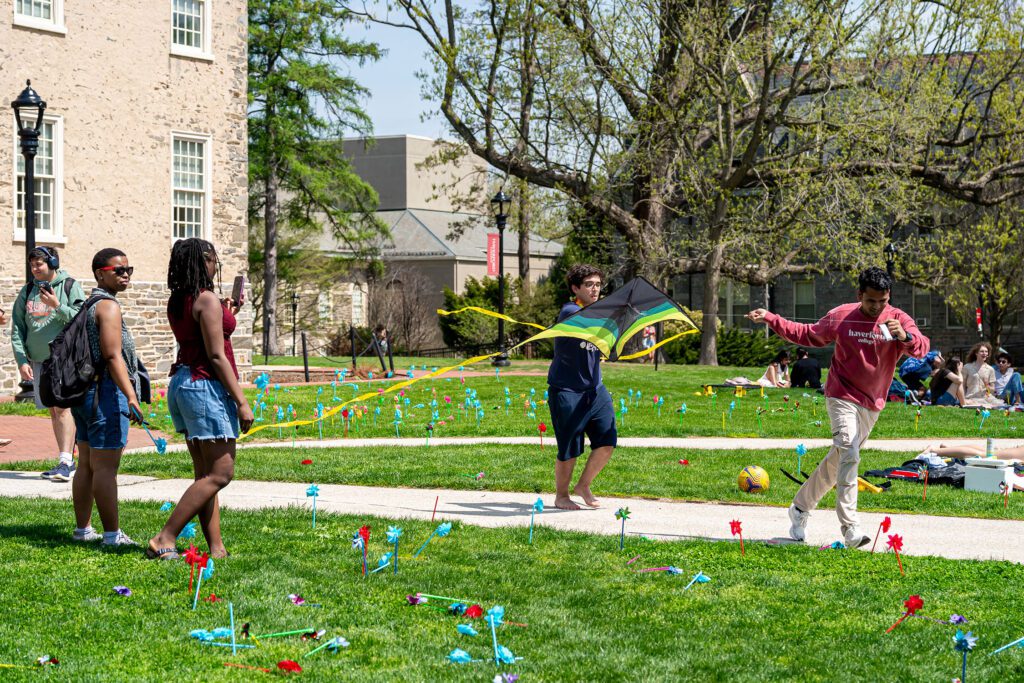 Students gather on the lawn as one runs with a black kite trailing long yellow and green ribbons across a field dotted with bright pinwheels.