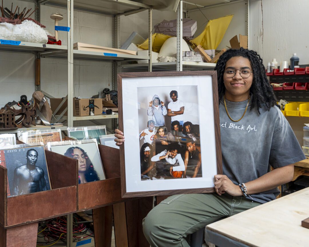 Ella Mbanefo ’26 sits in a studio holding a framed photograph, with shelves of art materials and bins of vinyl records behind her.