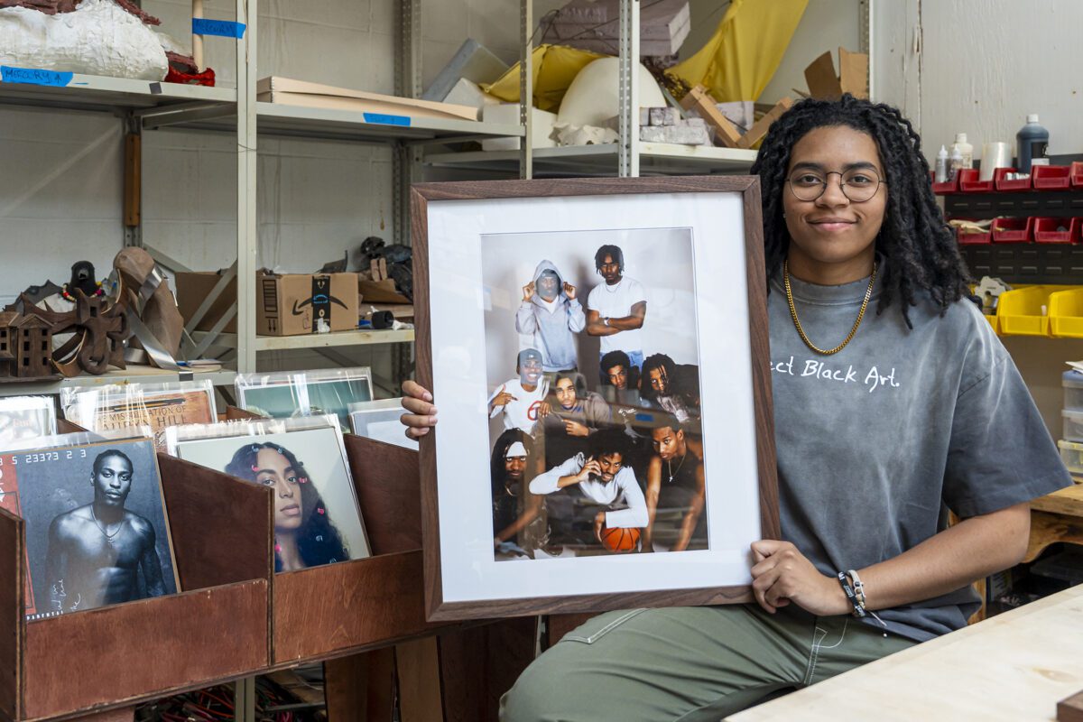 Ella Mbanefo ’26 sits in a studio holding a framed photograph, with shelves of art materials and bins of vinyl records behind her.