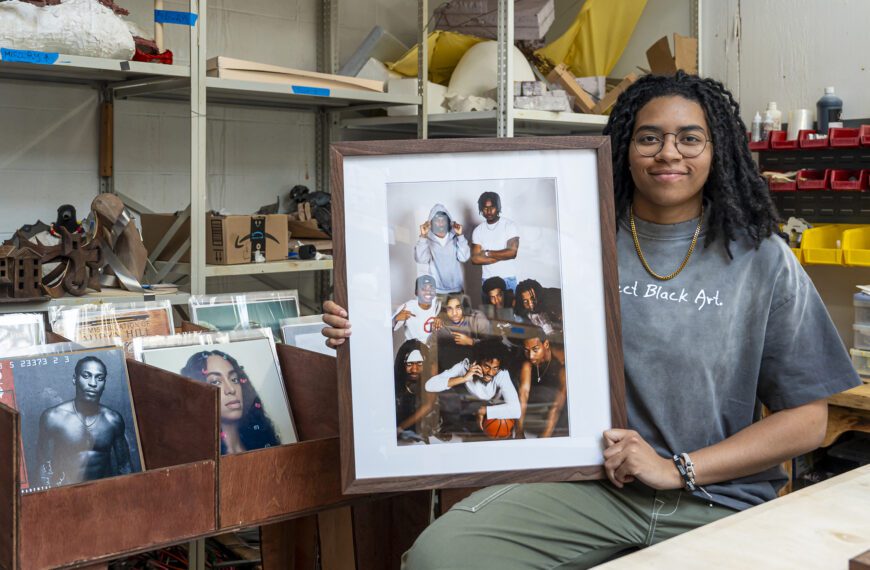 Ella Mbanefo ’26 sits in a studio holding a framed photograph, with shelves of art materials and bins of vinyl records behind her.
