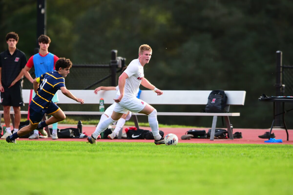 Andrew Fritz ’26 dribbles the ball upfield during a men’s soccer match as an opposing player closes in and teammates watch from the sideline.