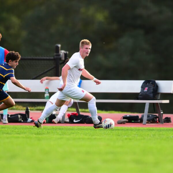 Andrew Fritz ’26 dribbles the ball upfield during a men’s soccer match as an opposing player closes in and teammates watch from the sideline.