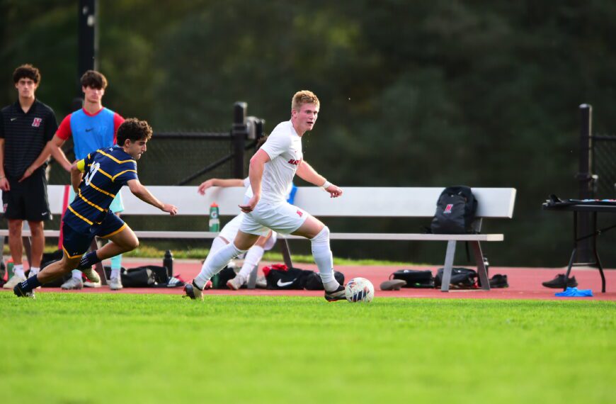 Andrew Fritz ’26 dribbles the ball upfield during a men’s soccer match as an opposing player closes in and teammates watch from the sideline.