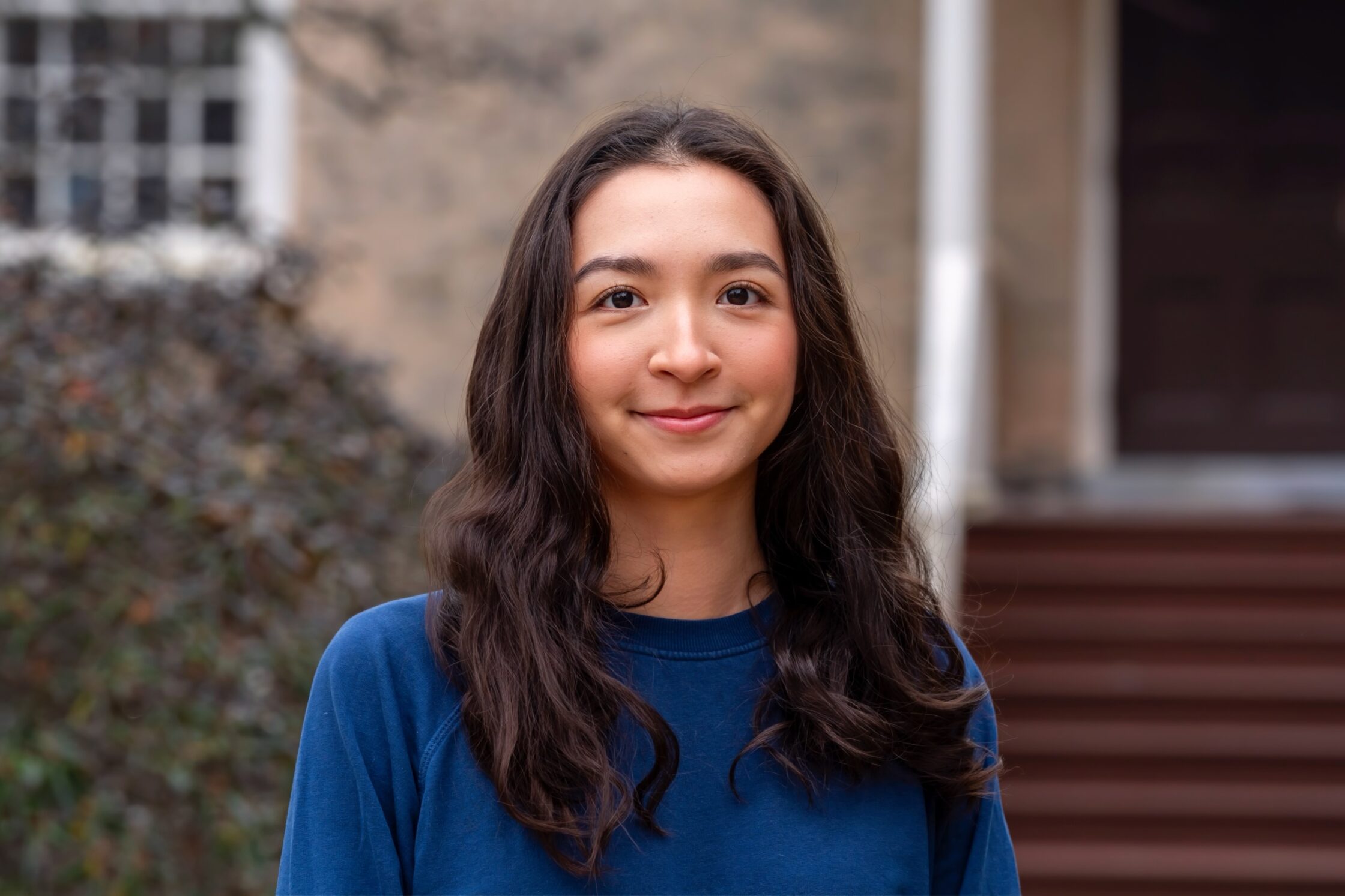 Alia Abulgazieva ’26 stands outdoors on campus in a blue top, smiling softly with a building entrance and shrubs blurred in the background.