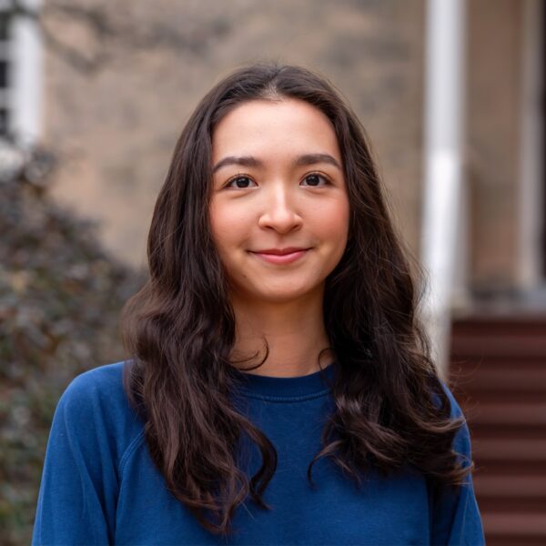 Alia Abulgazieva ’26 stands outdoors on campus in a blue top, smiling softly with a building entrance and shrubs blurred in the background.