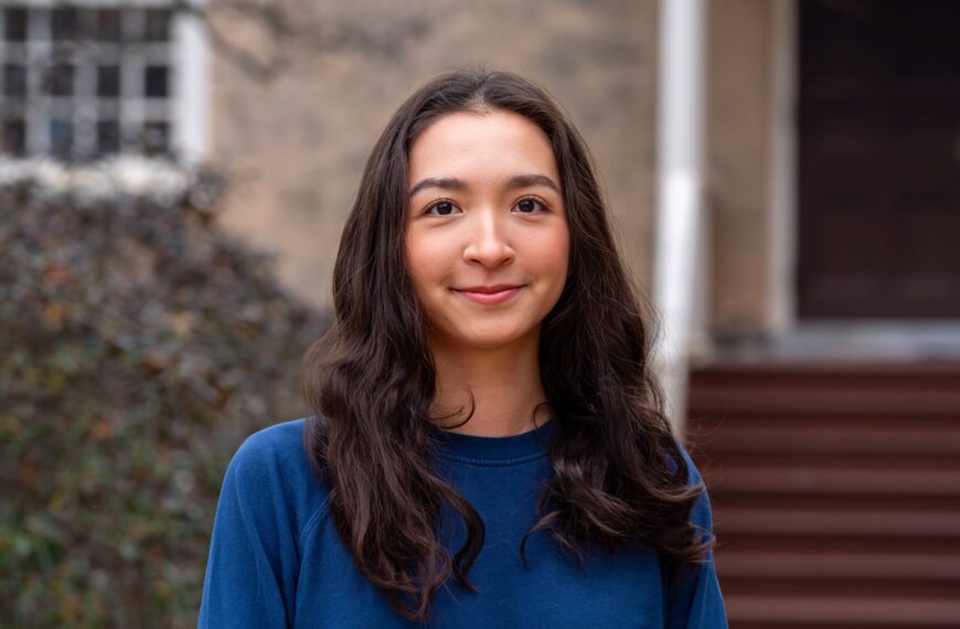 Alia Abulgazieva ’26 stands outdoors on campus in a blue top, smiling softly with a building entrance and shrubs blurred in the background.