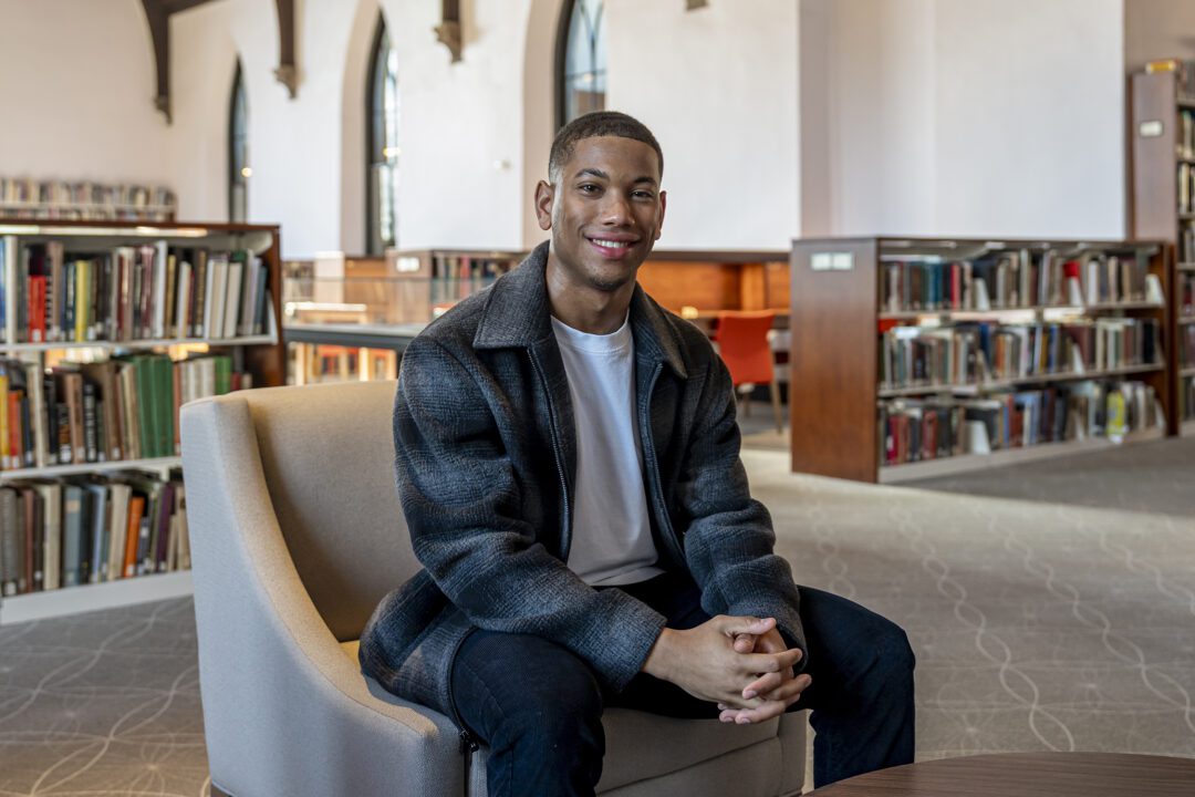Jackson Juzang ’26 sits smiling in a chair inside a campus library, with tall arched windows and bookshelves in the background.