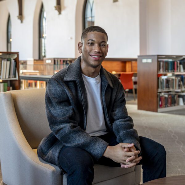 Jackson Juzang ’26 sits smiling in a chair inside a campus library, with tall arched windows and bookshelves in the background.