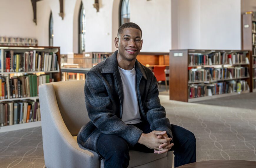 Jackson Juzang ’26 sits smiling in a chair inside a campus library, with tall arched windows and bookshelves in the background.