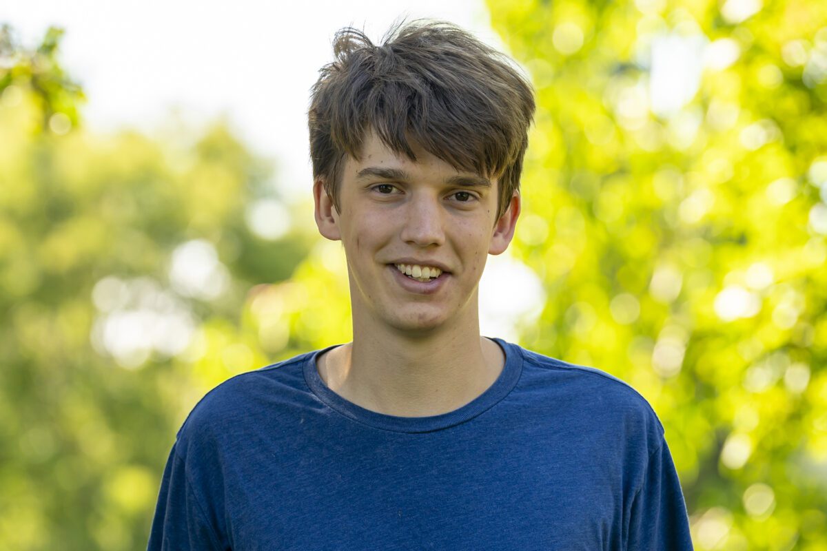 Oliver Wilson ’26 smiles outdoors on campus, wearing a blue T-shirt with sunlit green trees softly blurred in the background.