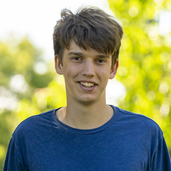 Oliver Wilson ’26 smiles outdoors on campus, wearing a blue T-shirt with sunlit green trees softly blurred in the background.
