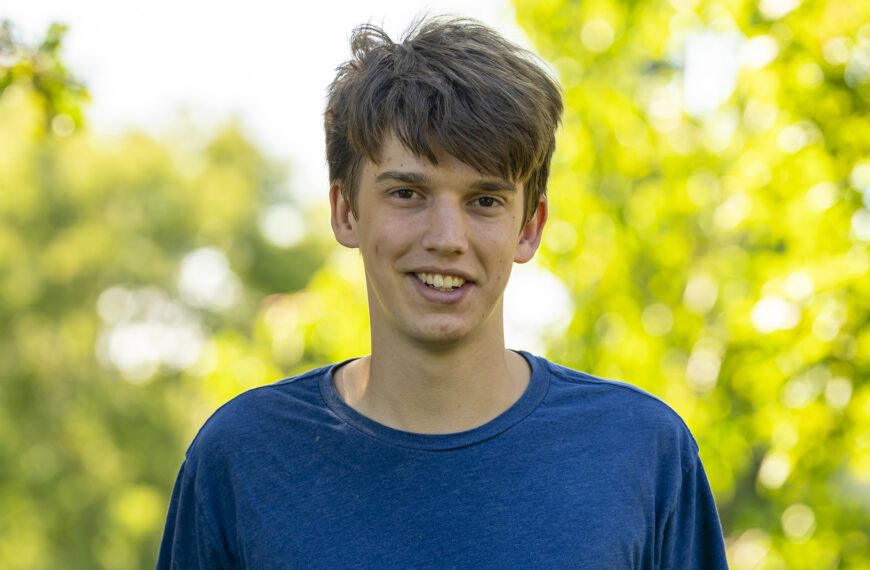 Oliver Wilson ’26 smiles outdoors on campus, wearing a blue T-shirt with sunlit green trees softly blurred in the background.