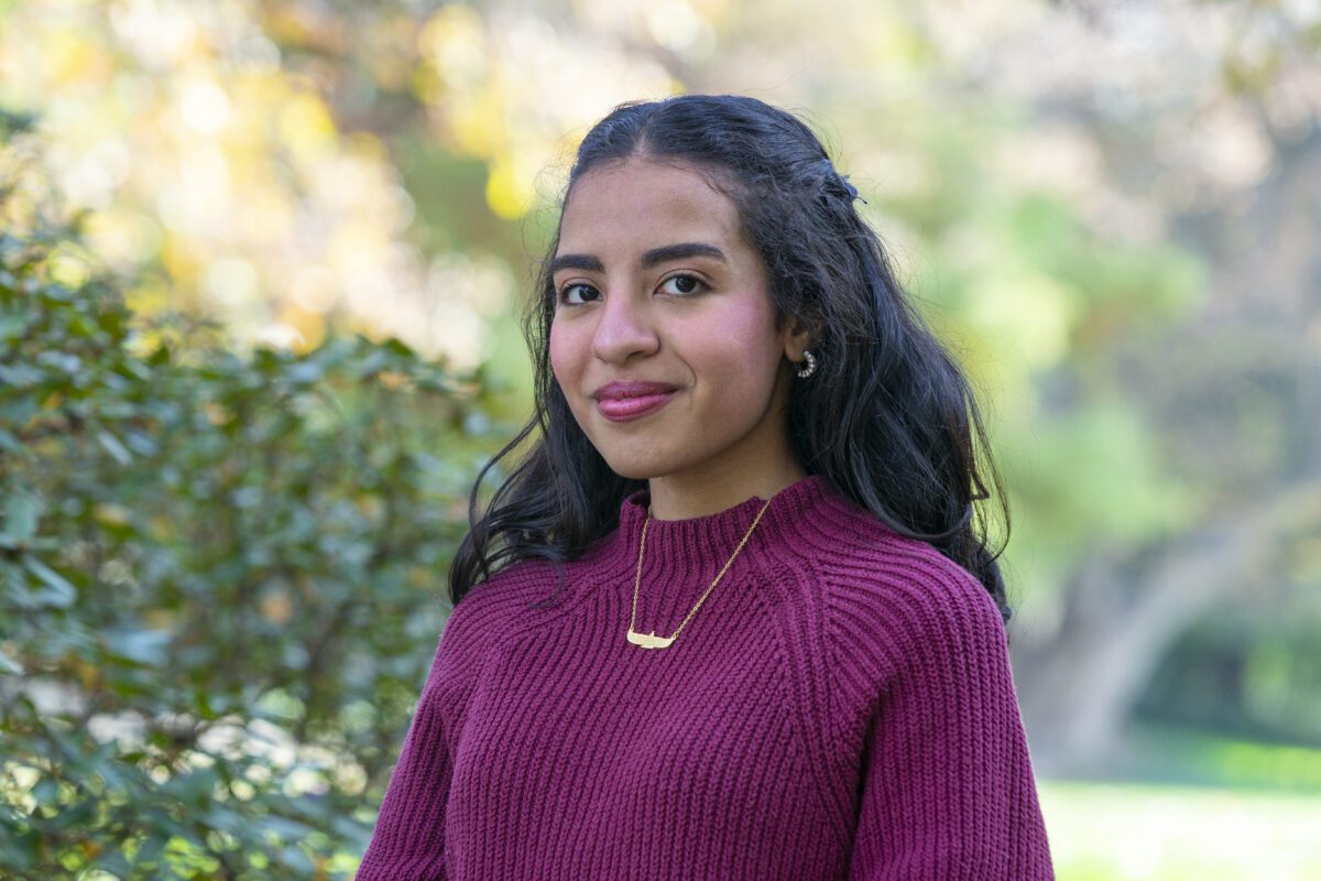 Nayera Hasan ’26 stands outdoors on campus in a burgundy sweater and gold necklace, with trees and greenery softly blurred in the background.